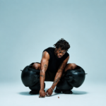 Kwes Darko crouching low in a striking press shot, wearing a black sleeveless top and sculptural oversized black leather trousers, with tattoos visible on both arms, against a pale blue studio background