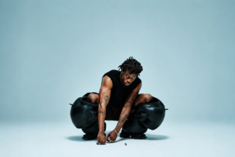 Kwes Darko crouching low in a striking press shot, wearing a black sleeveless top and sculptural oversized black leather trousers, with tattoos visible on both arms, against a pale blue studio background