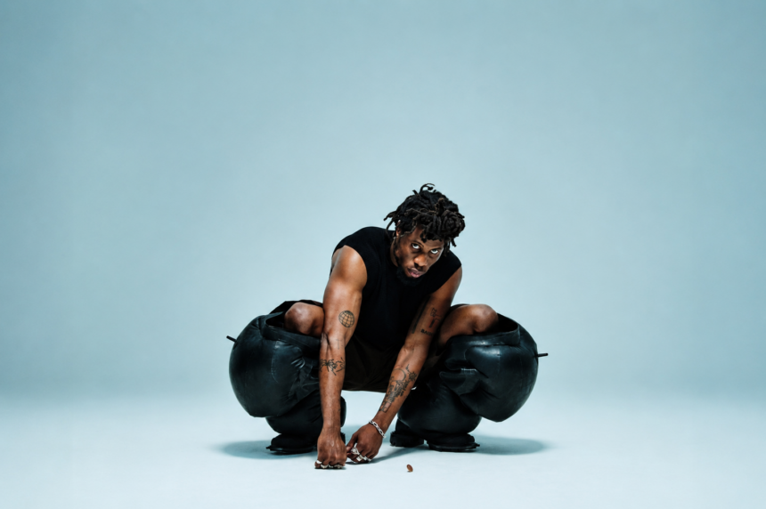 Kwes Darko crouching low in a striking press shot, wearing a black sleeveless top and sculptural oversized black leather trousers, with tattoos visible on both arms, against a pale blue studio background