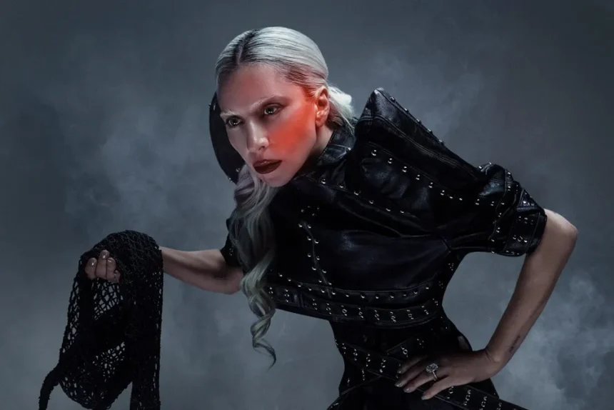 Lady Gaga in a studded black leather outfit, leaning forward dramatically against a smoky grey backdrop with red lighting.
