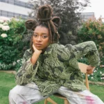 Little Simz seated outdoors in a garden, wearing a green snake-print top and tie-dye trousers, with her locs styled in an updo adorned with gold rings.