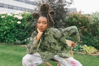 Little Simz seated outdoors in a garden, wearing a green snake-print top and tie-dye trousers, with her locs styled in an updo adorned with gold rings.