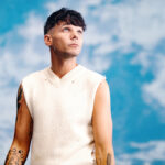 Louis Tomlinson gazing upward against a blue cloudy backdrop, wearing a cream sleeveless knit vest, with tattooed arms LateTown