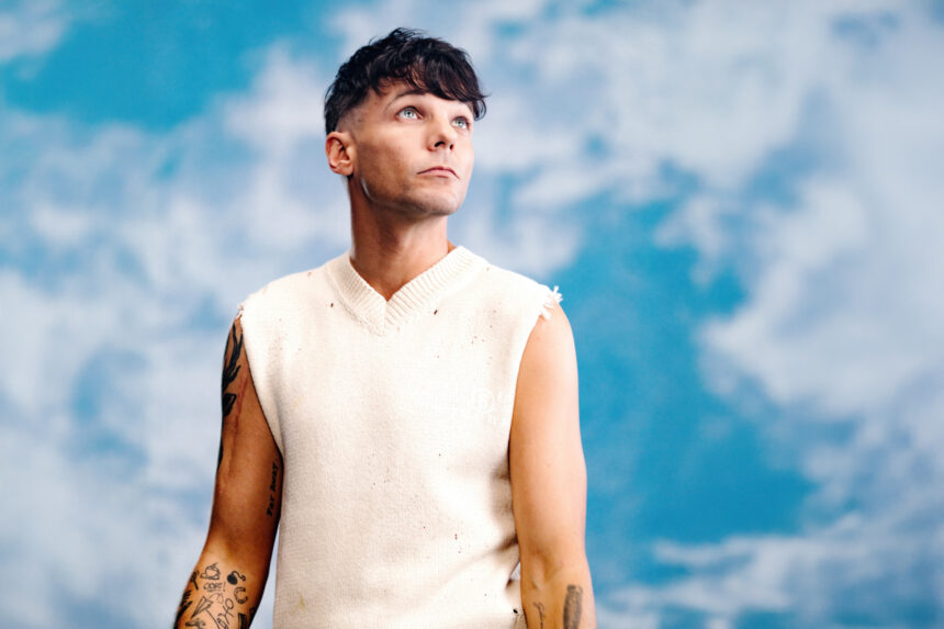 Louis Tomlinson gazing upward against a blue cloudy backdrop, wearing a cream sleeveless knit vest, with tattooed arms LateTown