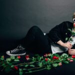 Man in white cat-eye sunglasses reclining on a dark floor surrounded by red roses, holding one rose to his lips.