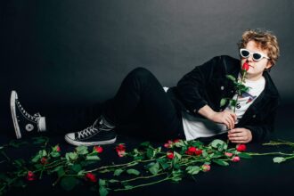 Man in white cat-eye sunglasses reclining on a dark floor surrounded by red roses, holding one rose to his lips.