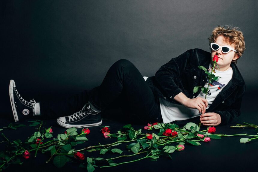 Man in white cat-eye sunglasses reclining on a dark floor surrounded by red roses, holding one rose to his lips.
