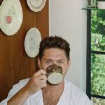 Niall Horan sipping from a speckled ceramic mug in a cozy room with decorative plates on a wooden door and a green garden visible through the window.