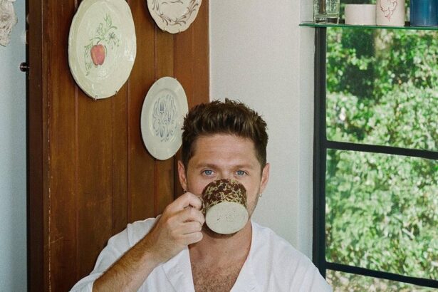 Niall Horan sipping from a speckled ceramic mug in a cozy room with decorative plates on a wooden door and a green garden visible through the window.
