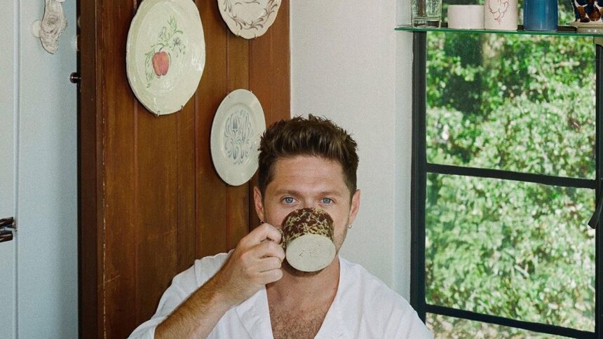 Niall Horan sipping from a speckled ceramic mug in a cozy room with decorative plates on a wooden door and a green garden visible through the window.