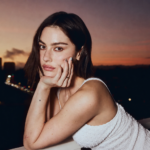 Portrait of Gracie Abrams leaning on a balcony at sunset with a city skyline in the background.