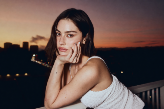 Portrait of Gracie Abrams leaning on a balcony at sunset with a city skyline in the background.