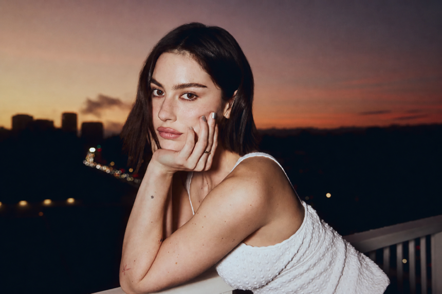 Portrait of Gracie Abrams leaning on a balcony at sunset with a city skyline in the background.