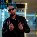 `ROMAN CIGI poses in a black leather jacket and sunglasses inside an airport terminal, in a press photo for his 'HEAVENUS' remix with Brosso.`