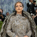 Rihanna at the 2026 Met Gala wearing a dramatic silver crystal-embellished Maison Margiela couture gown with a sculptural metallic cape, posing confidently on the red carpet as photographers capture the moment.