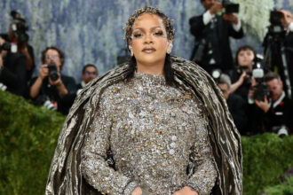 Rihanna at the 2026 Met Gala wearing a dramatic silver crystal-embellished Maison Margiela couture gown with a sculptural metallic cape, posing confidently on the red carpet as photographers capture the moment.
