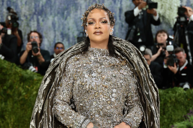 Rihanna at the 2026 Met Gala wearing a dramatic silver crystal-embellished Maison Margiela couture gown with a sculptural metallic cape, posing confidently on the red carpet as photographers capture the moment.