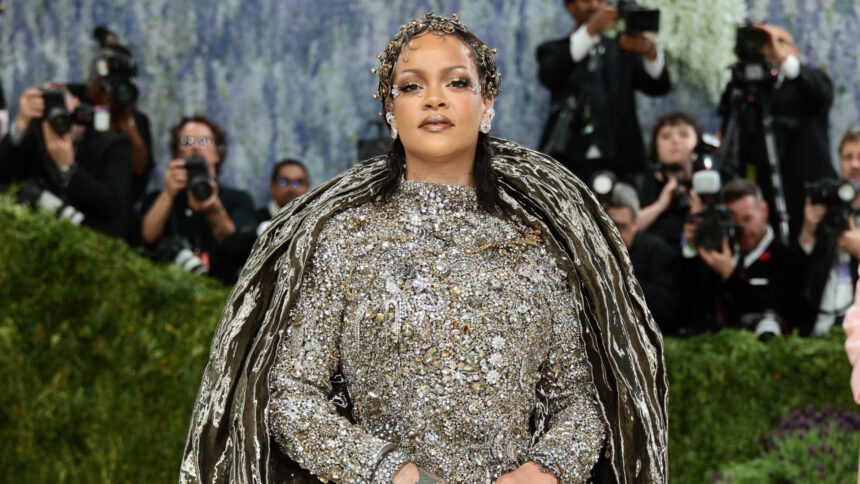 Rihanna at the 2026 Met Gala wearing a dramatic silver crystal-embellished Maison Margiela couture gown with a sculptural metallic cape, posing confidently on the red carpet as photographers capture the moment.