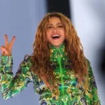 Shakira smiles broadly and flashes a peace sign on stage at her free concert in Rio de Janeiro, wearing a green, blue, and gold embroidered sequined outfit, with her long curly hair flowing freely.