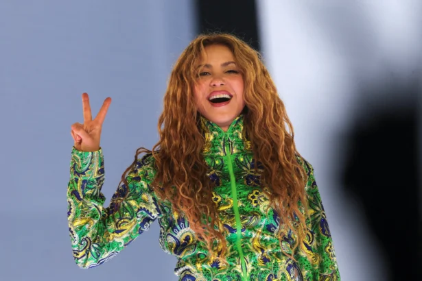 Shakira smiles broadly and flashes a peace sign on stage at her free concert in Rio de Janeiro, wearing a green, blue, and gold embroidered sequined outfit, with her long curly hair flowing freely.