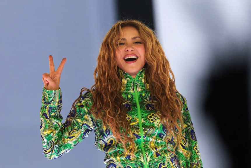 Shakira smiles broadly and flashes a peace sign on stage at her free concert in Rio de Janeiro, wearing a green, blue, and gold embroidered sequined outfit, with her long curly hair flowing freely.