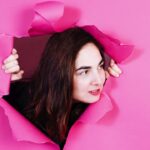 Shari Page peering through a torn hole in a bright pink paper backdrop, hands gripping the torn edges, with long dark hair and a subtle smile, shot in a playful and dynamic style