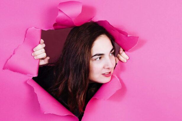 Shari Page peering through a torn hole in a bright pink paper backdrop, hands gripping the torn edges, with long dark hair and a subtle smile, shot in a playful and dynamic style