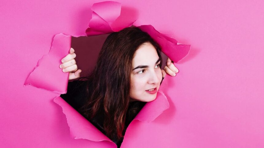 Shari Page peering through a torn hole in a bright pink paper backdrop, hands gripping the torn edges, with long dark hair and a subtle smile, shot in a playful and dynamic style