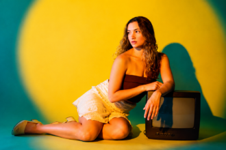Sophia Bavishi poses beside a vintage TV against a yellow-green backdrop in a press photo for her 2026 single 'FEEL IT.'
