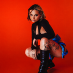 Talia Mar in her official press photo for her 2026 UK headline tour, crouching in a black cutout long-sleeve top, blue mini skirt and knee-high black leather boots, with floral tattoos on her thighs, against a vivid red studio background