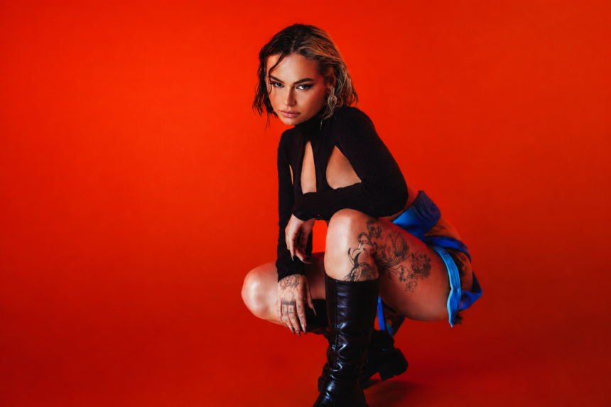 Talia Mar in her official press photo for her 2026 UK headline tour, crouching in a black cutout long-sleeve top, blue mini skirt and knee-high black leather boots, with floral tattoos on her thighs, against a vivid red studio background