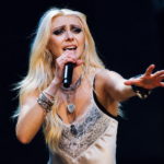 `Taylor Momsen performs live with The Pretty Reckless, singing into a microphone on stage ahead of their fifth album 'Dear God' out June 26, 2026.`