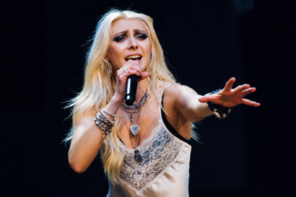 `Taylor Momsen performs live with The Pretty Reckless, singing into a microphone on stage ahead of their fifth album 'Dear God' out June 26, 2026.`
