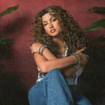 Tori Kelly seated against a red wall, hugging her knees, with an acoustic guitar and tropical plants nearby.