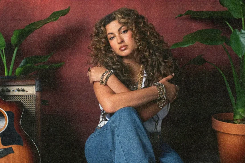 Tori Kelly seated against a red wall, hugging her knees, with an acoustic guitar and tropical plants nearby.
