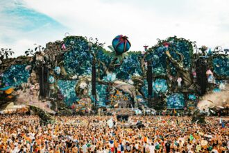 A massive crowd at Tomorrowland music festival in front of an elaborate fantasy-themed main stage decorated with giant flowers, trees, and mushrooms.