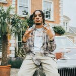 AJ Tracey sits on the hood of a classic Buick outside a brick mansion, wearing a plaid shirt, white tee, cargo pants, and a diamond chain, throwing up hand signs toward the camera.