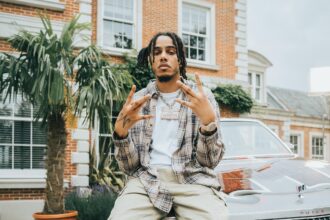 AJ Tracey sits on the hood of a classic Buick outside a brick mansion, wearing a plaid shirt, white tee, cargo pants, and a diamond chain, throwing up hand signs toward the camera.
