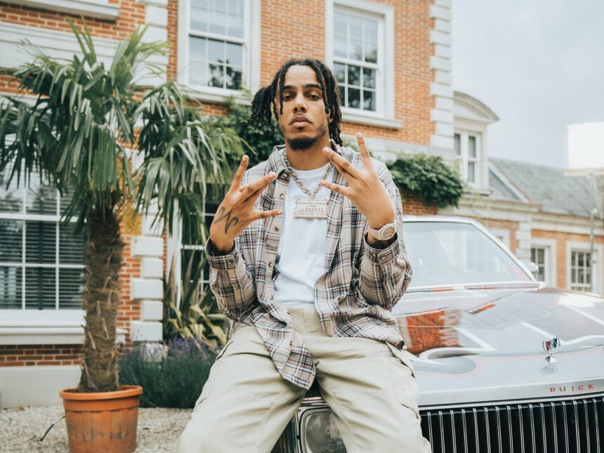AJ Tracey sits on the hood of a classic Buick outside a brick mansion, wearing a plaid shirt, white tee, cargo pants, and a diamond chain, throwing up hand signs toward the camera.