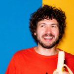 Baauer smiling in a vibrant studio portrait, wearing a bright orange shirt and holding a peeled banana against a bold blue-and-yellow background LateTown.com