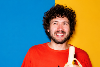 Baauer smiling in a vibrant studio portrait, wearing a bright orange shirt and holding a peeled banana against a bold blue-and-yellow background LateTown.com
