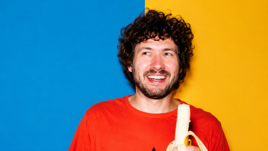 Baauer smiling in a vibrant studio portrait, wearing a bright orange shirt and holding a peeled banana against a bold blue-and-yellow background LateTown.com