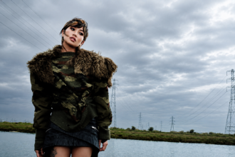 Beabadoobee poses by a riverside beneath dramatic storm clouds in a camouflage sweater and fur-trimmed jacket.