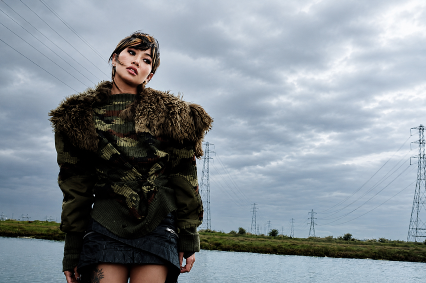 Beabadoobee poses by a riverside beneath dramatic storm clouds in a camouflage sweater and fur-trimmed jacket.