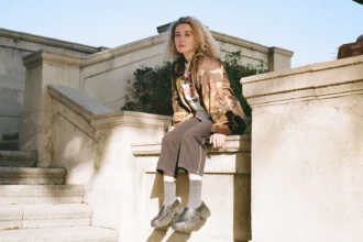 Blondshell seated on stone steps wearing a camo jacket, grey wide-leg pants, ribbed socks, and dark platform clogs.