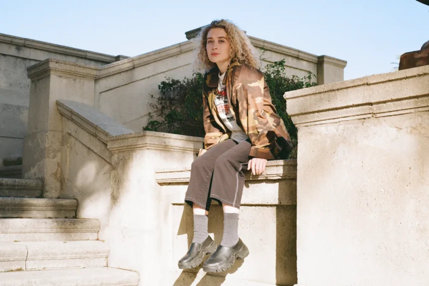 Blondshell seated on stone steps wearing a camo jacket, grey wide-leg pants, ribbed socks, and dark platform clogs.