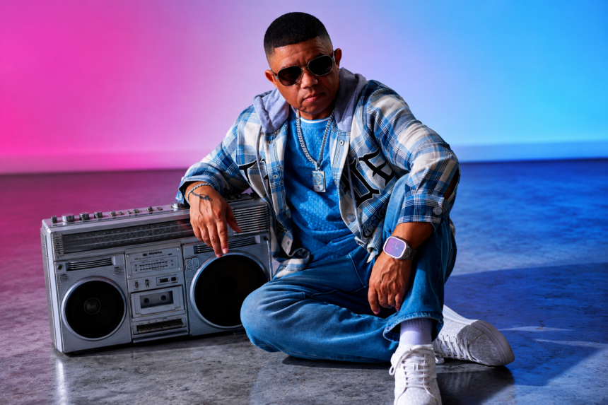 DJ Matrix seated on the floor next to a vintage boombox, wearing a blue plaid flannel shirt, jeans, silver chain, and aviator sunglasses in a studio setting