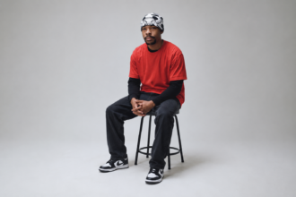 Evilgiane wearing a black and white patterned beanie and red t-shirt, photographed against a grey studio background
