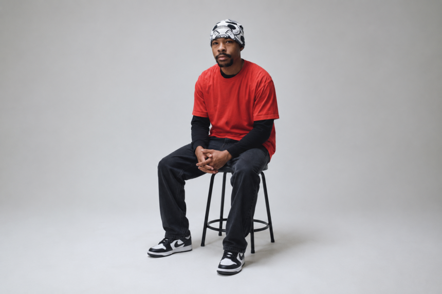 Evilgiane wearing a black and white patterned beanie and red t-shirt, photographed against a grey studio background