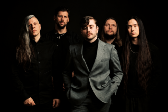 Imminence band press photo 2026 — five members posed against a dark background, with frontman Eddie Berg centered in a grey suit and black turtleneck, flanked by bandmates dressed in black